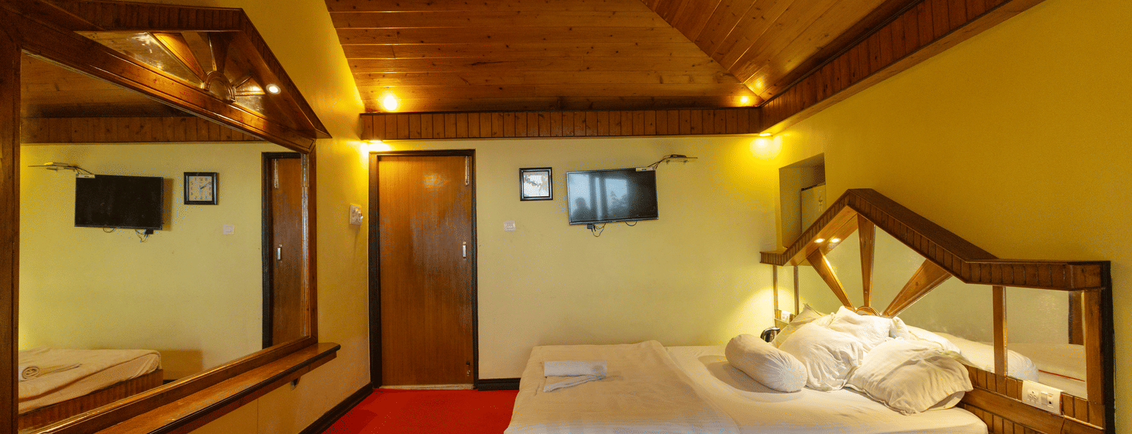 A bedroom with a king size bed, a wooden ceiling, a large mirror, a bedside table, and a wall mounted TV at Hotel Seagull Digha.
